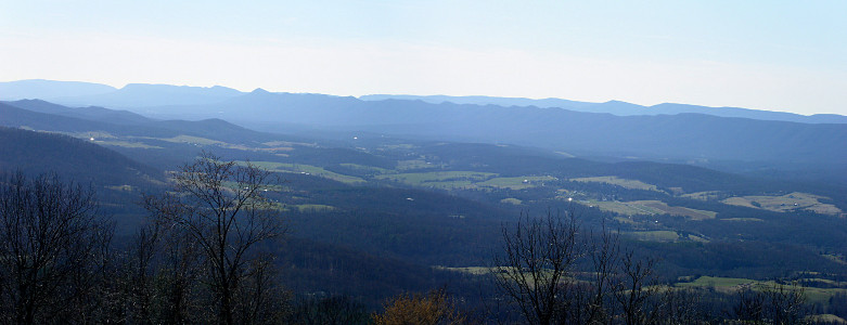 [Two photos stitched together showing an expanse of valley with some patches of green grass and many other patches of trees. There is a blue haze across much of the image, especially in the distance along the smooth-top mountains. There are a few leafless trees in the foreground which are sharp in color and contrast.]